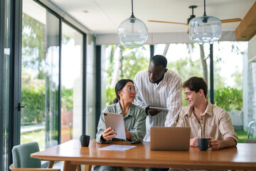 Three diverse young adults focus on a project, using tablets and laptops in a well-lit office, embodying teamwork and technology integration in a corporate setting.