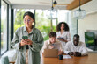 © Dorde - A joyful Asian businesswoman focused on her smartphone while colleagues engage in work behind her in a sunlit modern office space, reflecting the dynamic of today's diverse professional environments.