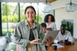 © Dorde - A content Asian young adult businesswoman enjoys her work on a laptop in a bright, contemporary office setting, with diverse colleagues focused on their tasks in the background.