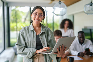 Wall Mural - A joyful Asian young adult businesswoman uses her laptop while standing in a brightly lit modern office, with diverse colleagues focused on work in the background.