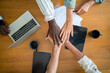© Dorde - A multiracial group of young adult business executives enthusiastically joins hands over a wooden table, surrounded by laptops and documents, signaling a unifying project triumph in a modern workspace