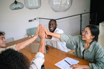 Wall Mural - Enthusiastic diverse young adults in professional attire engage in a team high five, expressing business success in a bright office environment, conveying achievement and unity.