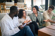 © Dorde - A group of diverse young adults engages in a discussion with a tablet, dressed in business casual attire, in a brightly lit modern office lounge area.