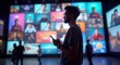© Vadym - Man uses phone in front of digital wall. Multiple screens show various video content. Wall represents social media marketing visuals. Young man in casual wear looks at device. Stands near digital