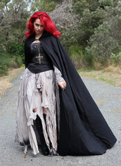 Wall Mural - full length portrait of beautiful red haired female model figure, wearing gothic fantasy costume with torn ballgown wedding dress, black corset, leather boots.  walking pose forest scenery background.