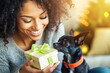© L.S. - Smiling woman with curly hair holding a gift box with a green ribbon, sitting next to a black Chihuahua with a red collar, in a cozy holiday setting.