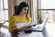 © fizkes - Focused Indian freelancer woman doing accounting paperwork at home, sitting at table with calculator and laptop, reviewing legal financial documents, bills, checking paper reports, invoices