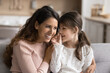 © fizkes - Preschooler 6s daughter sharing, whispering secrets to young loving mother, sit together on couch at home. Trust, closeness, trustworthy relationships between parent and child, family ties, close up