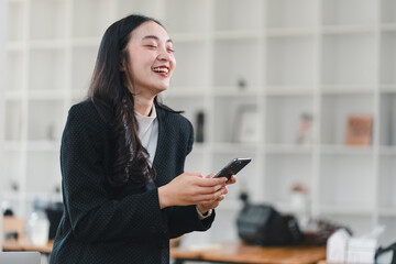 Wall Mural - joyful woman in black blazer smiles while using her smartphone in modern office. Her laughter adds lively touch to professional setting, showcasing moment of happiness