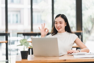 Wall Mural - Smiling woman in light shirt giving okay gesture while working on laptop in modern office. She exudes confidence and positivity