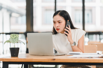 Wall Mural - Focused woman working on laptop while talking on phone, showing concentration and determination in modern office space