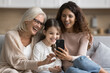 © fizkes - Multi-generational family using smartphone at home, sit on sofa look at device screen, laughing, watch funny online content, watching internet videos, making videocall using modern mobile application