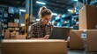 © Karina - Young woman working on a laptop in a busy warehouse setting