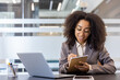 © Tetiana - Concentrated and smiling young African-American woman sitting in the office at the desk with a laptop and taking notes in a notebook, listening to a lecture and studying remotely