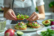 © Інна Мельник - Close-up photo of hands placing avocado on toast, next to a plate of vegetables and red onion. A woman in an apron sits at a table in a white kitchen.