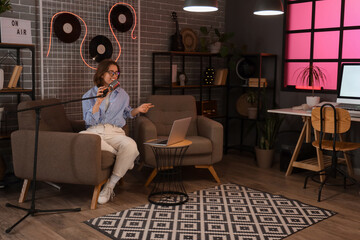 Wall Mural - Young woman with microphone and laptop recording podcast in studio