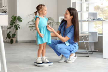 Wall Mural - Female pediatrician with cute little girl measuring weight on scales in clinic