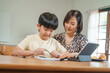 © NanSan - A mother teaches her son to study online at home, using a digital tablet for learning. They sit together at a table with snacks, promoting homeschooling and family bonding.