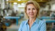 © Nadin Faust - A confident woman in a blue shirt stands with her arms crossed, exuding leadership in a bustling factory filled with machinery and equipment during daylight hours