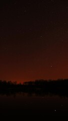 Wall Mural - Real Colorful Night Stars Above Lake River. Natural Starry Sky Background Backdrop. Time Lapse. Vertical Shot. Glowing Stars Reflected In Lake River Water. Natural Starry Sky Background Backdrop