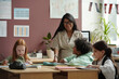 © pressmaster - Young cheerful teacher standing by table in front of several intercultural schoolkids and looking at one of them while explaining rules