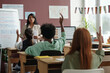 © pressmaster - Rear view of row of clever intercultural schoolkids raising hands while sitting by desks and listening to teacher at lesson
