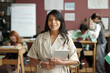 © pressmaster - Young smiling female teacher with tablet in hands looking at camera while standing in classroom at break against schoolkids