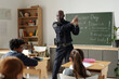 © pressmaster - Confident security guard in black uniform showing kids how to shoot gun while standing by blackboard on career day