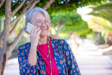 Wall Mural - Cheerful senior woman in public park while using mobile phone, caucasian elderly modern lady enjoying tech and social