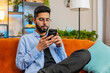 © Andrii Iemelianenko - Happy young man in casual clothes thinking and text messaging on smartphone. Indian guy sitting on sofa and using cellphone in living room at home. Male is searching information on browser internet.