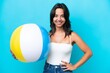 © luismolinero - Young hispanic woman holding beach ball isolated on blue background posing with arms at hip and smiling