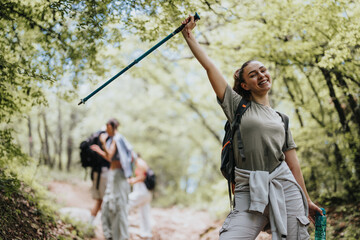 Wall Mural - Happy female hiker with friends on a forest trail, joyfully raising her trekking pole while enjoying nature.