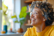 © Inga Bulgakova - African American joyful senior woman with curly gray hair and glasses wears a yellow sweater, smiling warmly in a bright indoor setting with plants and a blurred cozy background.