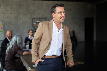 Fototapete - A man in a beige blazer stands confidently while engaging in a business meeting. Behind him, a diverse group collaborates at a large table, sharing thoughts in a stylish, contemporary office.
