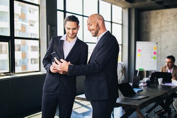 Fototapete - Two men in suits review documents on a device while collaborating in a stylish office. Natural light brightens the space, highlighting their engaged conversation and teamwork.