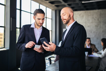 Fototapete - Two professionals engage in a discussion about project updates while reviewing data on a tablet. The office features large windows and a collaborative atmosphere with colleagues.