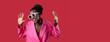 © Prostock-studio - A black woman dressed in a bright pink blazer and oversized sunglasses is playfully posing with raised hands, showcasing a vibrant and cheerful attitude against a bold red backdrop.