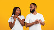 © Prostock-studio - Privacy Concept. Portrait of black man trying to look at his girlfriend's smartphone, woman hiding and covering screen of her gadget, couple standing isolated over yellow studio background