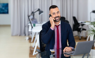 Poster - A cheerful man in a suit talks on the phone while sitting at a glass desk. A laptop and an espresso cup sit beside him in a sleek, well-lit office space filled with plants.