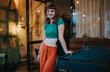 © qunica.com - A cheerful young woman with modern fashion stands in a cozy, stylish indoor cafe. The setting features warm lighting and comfortable seating, conveying a relaxed and welcoming atmosphere.