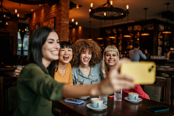 Wall Mural - A young group of people sitting around a cafe table, taking a group selfie and drinking coffee.