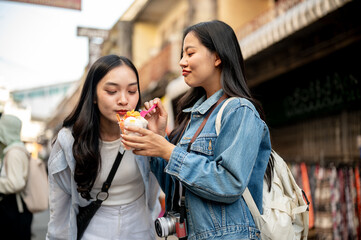 Two pretty, joyful Asian female tourists enjoy street food at a market in Thailand, eating ice cream