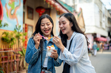 Two pretty, joyful Asian female tourists enjoy street food at a market in Thailand, eating ice cream