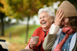 © Halfpoint - Portrait of a granddaughter and grandmother sitting on bench in autumn park, drinking coffee and laughing together.