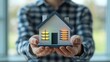 © Nathan - A man in a checkered shirt holds a model house with illuminated windows, symbolizing home ownership and security.