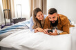 © djoronimo - Young happy couple is on vacation. Tourists arrived in hotel room with their suitcases. They are lying on bed and using smartphone.