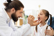 © sofiko14 - Caucasian male doctor examines young Caucasian girl's throat using a wooden tongue depressor. Girl sits calmly in medical office setting, showing a trustful patient-doctor interaction.