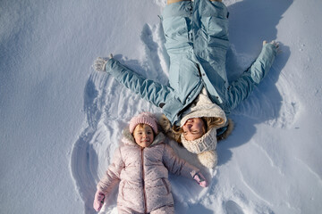 Wall Mural - Portrait of small girl and mom making snow angels, enjoying winter holiday in the mountains. Girl lying and playing in snow.