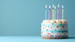 © COK House - A festive birthday cake with brown base, white stripes, yellow & white frosting, colorful spots and 7 lit candles on blue background exudes celebratory vibes.