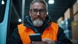 ©  Shomixer - A bearded man with glasses and a safety vest holds a smartphone inside a warehouse, illustrating safety, connectivity, and professional alertness.